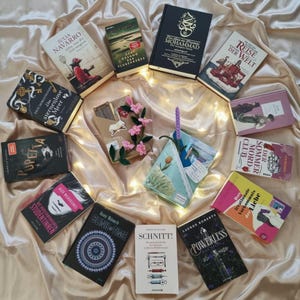 Könnte beinhalten: Eine Sammlung verschiedener Bücher, die kreisförmig auf einer drapierten Stoffoberfläche angeordnet sind. Die Bücher haben unterschiedliche Coverdesigns, darunter Titel wie 'Das sternenlose Meer', 'Powerless' und 'Schnitt', sowie ein kleines Deko-Objekt mit rosa Blüten.