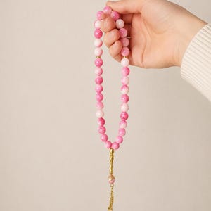 Könnte beinhalten: Eine rosa-weiße Perlenkette mit einer goldfarbenen Quaste und einem dekorativen Anhänger. Die Kette wird in einer Hand gehalten und zeigt die verschiedenen Rosa- und Weißtöne. Quaste und Anhänger verleihen einen Hauch von Eleganz.