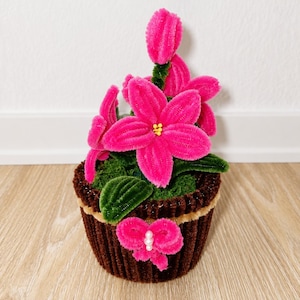 Könnte beinhalten: Ein handgefertigtes Blumenarrangement aus flauschigen Pfeifenreinigern mit leuchtend pinken Blütenblättern, gelben Mitten und dunkelgrünen Blättern. Die Blumen stecken in einem braunen, gerippten, tassenförmigen Sockel mit einer pinken Schleife.