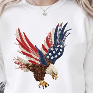 May include: White sweatshirt with an embroidered eagle design. The eagle's wings are patterned in red, white, and blue, with stars and stripes. The eagle has brown feathers and yellow beak and talons. The text at the bottom reads "HIGH QUALITY . 300DPI PNG . FILE".