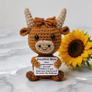 Op de afbeelding: Een gehaakte bruine koeienfiguur met bruine hoorns en een bruine snuit. De koe houdt een bord vast met "Positive Moo" en een inspirerende boodschap. Op de achtergrond staat een gele zonnebloem.