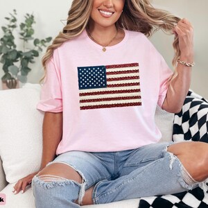 May include: Light pink t-shirt featuring a sequined American flag design. The flag has a blue square with white stars in the upper left corner and red and white stripes. The shirt is worn by a person.