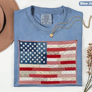 Könnte beinhalten: Ein blaues Jeans-T-Shirt mit einem Patchwork-Design der amerikanischen Flagge. Die Flagge zeigt ein blaues Sternenfeld und rot-weiße Streifen. Eine goldene Sternanhänger-Halskette liegt auf dem Shirt. Das Shirt ist mit XL-TG-EG gekennzeichnet.