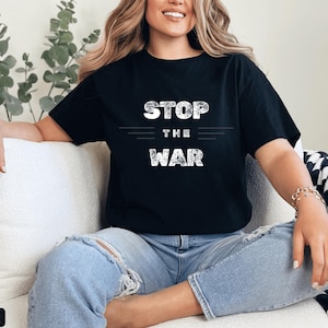 Pode incluir: Camiseta preta com o texto "STOP THE WAR" em branco, com fonte desgastada. A camisa é usada com jeans azuis. A imagem mostra uma pessoa sentada em um sofá branco com uma almofada xadrez em preto e branco.