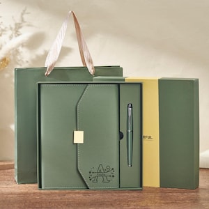 May include: A green gift set comprising a journal, pen, and gift bag. The journal has a gold clasp and an initial design. The pen is silver. The gift bag has a light brown handle. The set is displayed on a wooden surface.