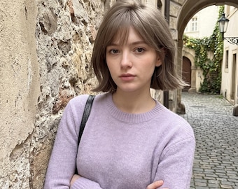 Könnte beinhalten: Ein langärmeliger Pullover mit Rundhalsausschnitt in einem sanften Lavendelton. Das Strickkleidungsstück verfügt über einen gerippten Halsausschnitt und Bündchen sowie eine texturierte, gewebte Optik. Ein schwarzer Schultertaschenriemen ist über der Schulter zu sehen.