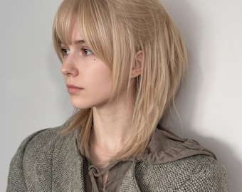 Könnte beinhalten: Eine schulterlange blonde Perücke mit geradem Pony und gestuften Spitzen. Das Haar hat einen hellen Aschblondton mit dezenten Highlights. Die Perücke ist mit einer weichen, natürlichen Textur gestylt und wird über einem braunen Kapuzenoberteil getragen.