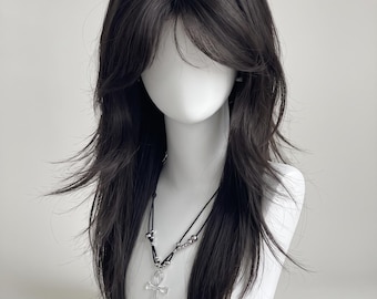 May include: A long, dark brown wig with layered, feathered ends and a center part, styled with soft volume around the crown. A black cord necklace with silver-toned beads and a clear, cross-shaped ankh pendant rests on the neck of the mannequin.