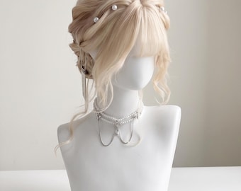 Könnte beinhalten: Eine platinblonde Perücke, die zu einer aufwendigen Hochsteckfrisur mit weichem Pony und kleinen weißen Perlenakzenten im Haar gestylt ist. Die Perücke wird auf einem weißen Schaufensterpuppenkopf mit einer mehrlagigen Perlen- und Silberkette präsentiert.