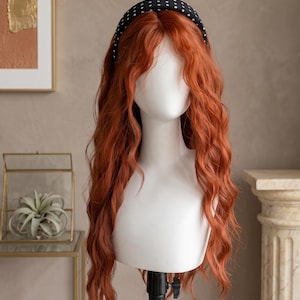 May include: A long, wavy, copper-colored wig styled with a center part and a black headband featuring a white polka dot pattern. The wig is displayed on a white mannequin head, showcasing vibrant orange-red hair with loose, flowing curls.