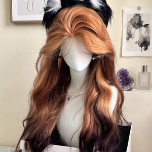 May include: A long, wavy wig featuring a gradient of ginger and copper tones transitioning into dark brown at the ends. The wig is styled with a center part and is adorned with a pair of black and white faux fur cat ears on a headband.