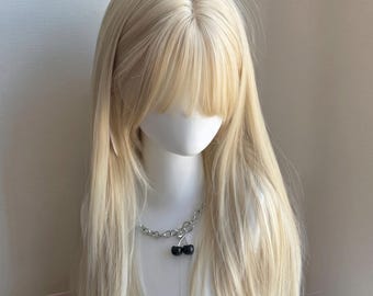 Könnte beinhalten: Eine lange, glatte, platinblonde Perücke mit Pony auf einem weißen Schaufensterpuppenkopf. Die Puppe trägt eine silberfarbene Gliederkette mit einem Anhänger, der aus zwei kleinen, runden, schwarzen, kirschförmigen Elementen besteht.