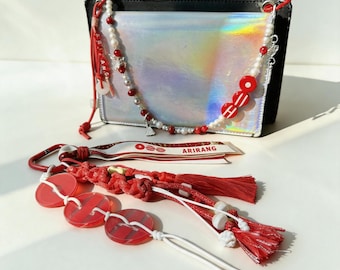 May include: A collection of red and white accessories, including a beaded necklace, keychains, and a ribbon with the word "ARIRANG". The items are arranged in front of a silver and black bag. The necklace has red and white beads.