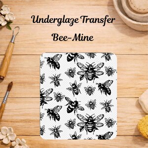 Op de afbeelding: Een rechthoekig onderglazuur transfervel met een herhalend zwart-wit patroon van diverse vliegende bijen. De tekst "Underglaze Transfer" en "Bee-Mine" is in het zwart gedrukt boven het vel op een licht houten oppervlak.