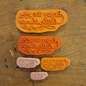 Op de afbeelding: Vijf oranje en lichtroze rubberen stempels van verschillende groottes, elk voorzien van de reliëftekst "Enjoy the little things" met een decoratieve bladergrafiek die de tekst in een cursieve stijl onderstreept en omlijst.