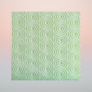 Pode incluir: Uma placa quadrada de cor verde menta claro texturizada com um padrão repetitivo de círculos concêntricos. A superfície é gravada com múltiplos anéis circulares de vários tamanhos que criam um design geométrico tátil no painel.