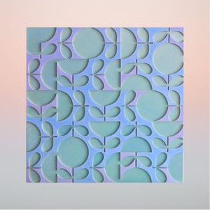 Op de afbeelding: Een vierkant wandpaneel met een herhalend geometrisch patroon van gestileerde bloemen en bladeren. Het ontwerp bestaat uit verhoogde lichtblauwe cirkelvormige en halfronde vormen tegen een vlakke, lichtpaarse achtergrond met een textuureffect.