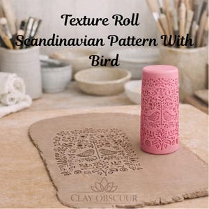 Op de afbeelding: Een roze cilindrische textuurrol met een gedetailleerd Scandinavisch patroon met vogels en bladeren. Het gereedschap ligt naast een plat stuk klei dat hetzelfde reliëfpatroon vertoont. De tekst luidt Texture Roll Scandinavian Pattern With Bird.