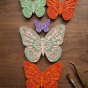 Op de afbeelding: Een verzameling van vijf gelaagde vlinderuitsparingen in mintgroen, oranje en paars, met ingewikkelde filigraanpatronen. De vlinders zijn in een groep gerangschikt naast een kleine verfkwast en een metalen pincet.