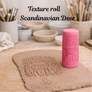 Op de afbeelding: Een roze cilindrische textuurroller met een Scandinavisch hertontwerp. Het gereedschap ligt naast een plat stuk bruine klei met een reliëfafdruk van hetzelfde hertpatroon. De tekst Texture roll Scandinavian Deer is duidelijk zichtbaar.