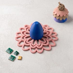 May include: A blue egg-shaped object sits atop a pink flower-shaped coaster. Nearby is a small, layered dome-shaped object with pink and beige petals and a textured top, alongside three small green square tiles and one small gold square tile.