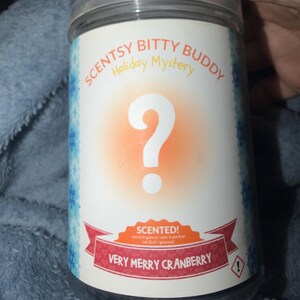 May include: A clear plastic jar with a white label featuring the text "SCENTSY BITTY BUDDY Holiday Mystery" and "VERY MERRY CRANBERRY". The label has a large question mark and a red banner that says "SCENTED!".