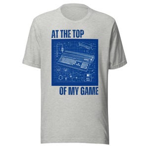 May include: A light gray t-shirt featuring a blue blueprint design of a vintage computer keyboard with the text "AT THE TOP OF MY GAME". The design includes the text "A500" and other technical details.