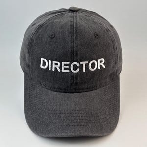May include: A charcoal grey baseball cap with a washed-out, distressed texture. The word DIRECTOR is embroidered in white capital letters across the front centre of the crown. The cap features a curved brim and six panels with stitched eyelets.
