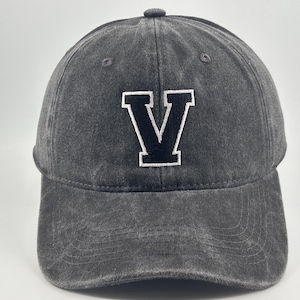 May include: A charcoal grey baseball cap with a distressed, washed-out fabric finish. The front panel features a large, bold letter V embroidered in black with a thick white outline. The cap has a curved brim and stitched eyelets on the crown.