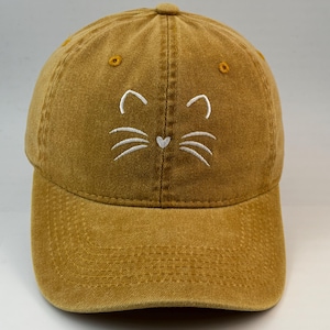 Op de afbeelding: Een mosterdgele baseballpet met een verweerde, verwassen afwerking. Het voorpaneel is voorzien van wit borduurwerk bestaande uit twee puntige kattenoren, drie snorharen aan elke kant en een klein hartje dat een neusje voorstelt.