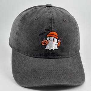 Puede incluir: Una gorra de béisbol gris carbón con un diseño bordado de un fantasma blanco que lleva un gorro naranja. El fantasma sostiene una pequeña calabaza naranja y un vaso naranja, con dos pequeños pájaros negros volando sobre su cabeza.