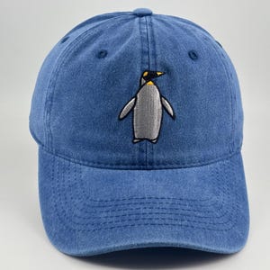 Puede incluir: Una gorra de béisbol azul con acabado desgastado que presenta un pingüino bordado en el panel frontal. El pingüino está representado en tonos gris, blanco y amarillo. La gorra tiene una visera curva con costuras visibles y un botón superior.