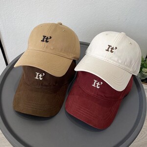 May include: Four baseball caps in tan, dark brown, white, and maroon. Each cap features a curved brim, a button on top, and the embroidered text "It'" on the front panel. The caps are stacked in two pairs, showcasing the varied solid colours.