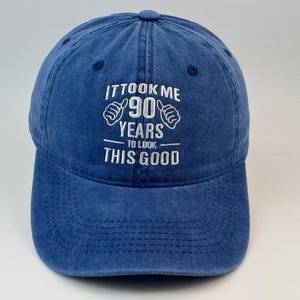 Op de afbeelding: Een blauwe honkbalpet met witte geborduurde tekst die luidt IT TOOK ME 90 YEARS TO LOOK THIS GOOD, inclusief twee duim-omhoog iconen aan weerszijden van het getal 90. De pet heeft een constructie van zes panelen met een gebogen klep en knoop.