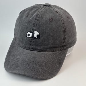 Può includere: Un cappellino da baseball grigio antracite con una finitura slavata in stile vintage. Il pannello frontale presenta una piccola toppa ricamata di un panda che dorme, realizzata con filo bianco e nero. Il cappello ha una visiera curva e un bottone.