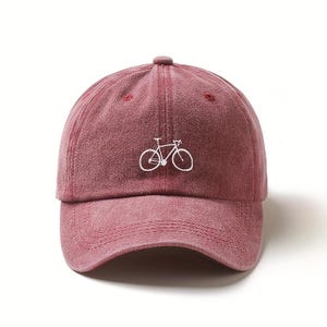 May include: A washed-out red baseball cap featuring a simple white embroidered outline of a road bicycle centered on the front panel. The cap has a curved brim, a button on top, and stitched eyelets for ventilation on the crown panels.