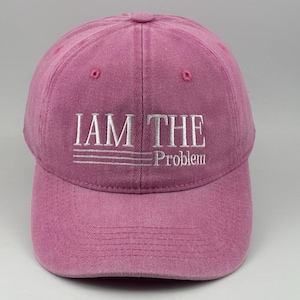 Peut inclure: Une casquette de baseball rose poudré avec une inscription brodée en blanc indiquant I AM THE Problem, accompagnée de trois lignes horizontales sous les deux premiers mots. La casquette présente une calotte structurée et une visière courbée.
