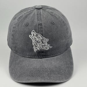 Può includere: Un cappellino da baseball grigio antracite con un ricamo bianco raffigurante una testa di lupo che ulula sul pannello frontale. Il cappellino presenta una struttura a sei pannelli con visiera curva, un bottone sulla parte superiore e tessuto.