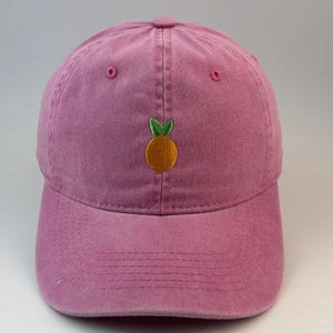 Pode incluir: Um boné de beisebol rosa empoeirado com um pequeno bordado centralizado de uma laranja com duas folhas verdes. O boné possui uma copa estruturada, aba curva e detalhes de costura em rosa, incluindo ilhós nos painéis superiores.