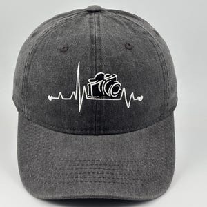 Puede incluir: Una gorra de béisbol gris carbón con bordado blanco en la parte delantera. El diseño muestra un patrón de línea de latido del corazón que incorpora un icono de cámara en el centro y pequeñas formas de corazón en los extremos.