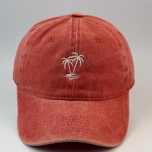 Könnte beinhalten: Eine verwaschene rote Baseballkappe mit einer weißen Stickerei von zwei Palmen auf der Vorderseite. Die Kappe hat einen gebogenen Schirm, eine Sechs-Panel-Konstruktion mit genähten Ösen und eine strukturierte Krone mit verwaschener Optik.