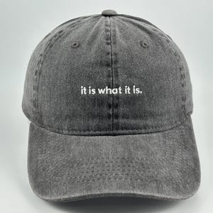 Pode incluir: Um boné de beisebol cinza-carvão com uma textura lavada e desgastada. O painel frontal apresenta o texto bordado em branco "it is what it is.". O boné tem uma aba curva, um botão no topo central e ilhós costurados para ventilação.