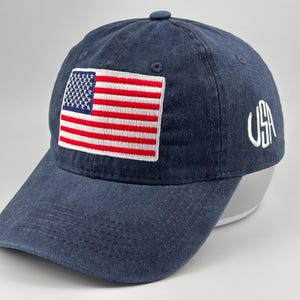 Puede incluir: Una gorra de béisbol azul marino con un parche bordado de la bandera estadounidense en el panel frontal. El lateral de la gorra muestra el texto USA bordado en blanco con una tipografía estilizada. La gorra tiene una visera curva y seis paneles.
