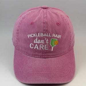 Könnte beinhalten: Eine staubrosafarbene Baseballkappe mit weißem Sticktext, der PICKLEBALL HAIR don't CARE lautet. Das Design enthält einen kleinen grünen Pickleball-Schläger mit einem gelben Ball-Symbol, das rechts neben dem Wort don't eingestickt ist.