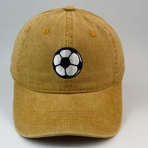 Puede incluir: Una gorra de béisbol de color amarillo mostaza con un acabado desgastado y lavado. En la parte central delantera de la gorra hay un gráfico bordado de un balón de fútbol clásico en blanco y negro. Incluye visera curva y ojales amarillos.