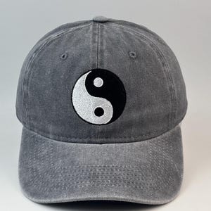 Könnte beinhalten: Eine anthrazitfarbene Baseballkappe mit verwaschener Stoffoptik. Auf der Vorderseite befindet sich ein kreisförmiger, gestickter Aufnäher mit einem schwarz-weißen Yin-Yang-Symbol, das mittig auf der Kappe platziert ist, inklusive genähter Ösen.