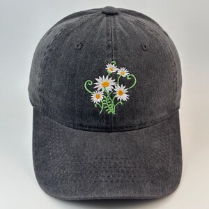May include: A charcoal grey baseball cap featuring an embroidered design of white daisies with yellow centres and green stems. The cap has a six-panel construction with a curved brim and a button on the top, showcasing a washed-out fabric texture.