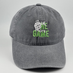May include: A charcoal grey baseball cap featuring an embroidered design on the front. The embroidery includes a white pickleball graphic and the green text "Just ONE more GAME" stacked in a stylised font with a button on the crown.