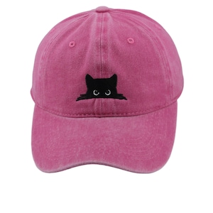 May include: A bright pink baseball cap with a distressed, washed-out finish. The front panel features an embroidered black cat graphic, showing only the head and ears of the cat peeking out, with two small white eyes visible in the centre.
