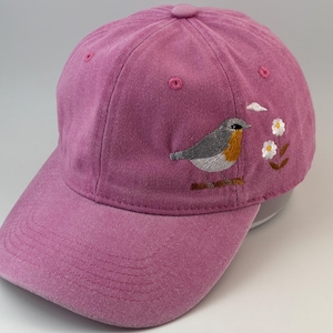 Puede incluir: Una gorra de béisbol de color rosa empolvado con un bordado de un pequeño pájaro gris y naranja junto a dos flores blancas con centro amarillo y una pequeña nube blanca. La gorra tiene una visera curva y ojales cosidos en la corona.
